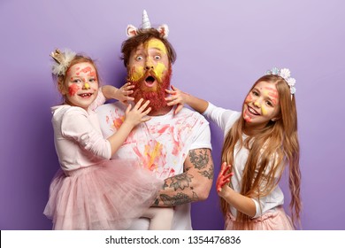 Funny two girls paint fathers beard with colorful paints, have fun together, celebrate holiday, wear festive apparel, have ginger hair, look alike, stand over purple background. Childhood concept - Powered by Shutterstock