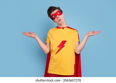 Funny uncertain male superhero in bright cape and mask shrugging and looking up on blue background in studio
