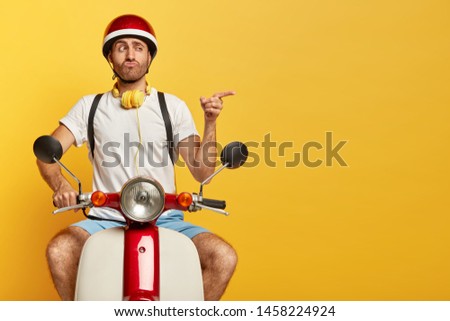 Funny unshaven guy in casual wear, drives on motorcycle, has good trip, enjoys freedom, points index finger on blank space over yellow wall, pouts lips. Professional driver bought new scooter