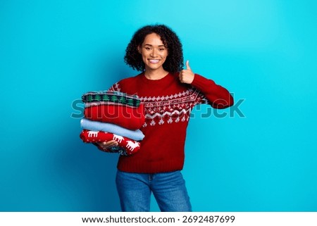 Funny and warm inspired caption about a joyful young multiethnic woman stacking cozy knits and giving thumbs up against blue background