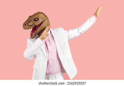 Funny weird guy in wacky animal mask having fun at crazy party. Eccentric man in white suit and silly ugly masquerade dinosaur mask dancing isolated on pink background
