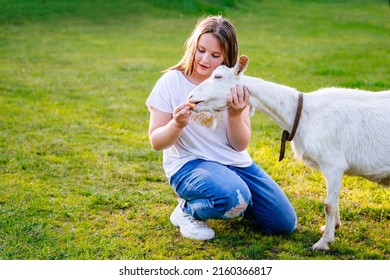 Funny white purebred goat on the goat farm. Agrotourism. Farm domestic animals. Summer sunny day in village. Happy teenager girl feeding goat on smallholding farm.Girl in garden with a goat.