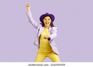Funny woman dancing against lilac background. Studio shot of happy beautiful young girl in purple hat, lilac jacket and yellow eyeglasses crying Yeehaw, dancing like a cowgirl and enjoying music
