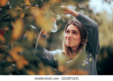 
Funny Woman Hanging Light-bulb Decorations In A Tree. Green Energy Concept Image With A Worried Person Unscrewing A Light Bulb
