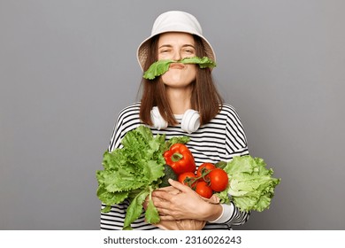 Funny woman holding vegetables isolated over gray background making mustache with green lettuce looking at camera, having fun, grimacing.