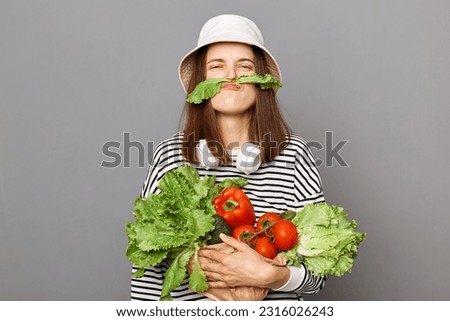 Funny woman holding vegetables isolated over gray background making mustache with green lettuce looking at camera, having fun, grimacing.
