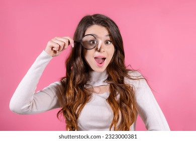 Funny woman looking through magnifying glass against pink background standing in white sweater. Searching information concept. 