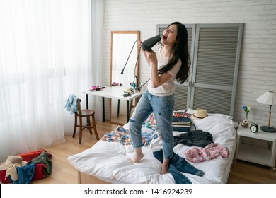 Funny Woman Singing In Hair Dryer At Home Standing On Messy Bed Full Of Clothes Dresses. Young Asian Girl Packing Suitcase In House Bedroom Having Fun With Tools Dancing Indoors In Day Time Sunny Day
