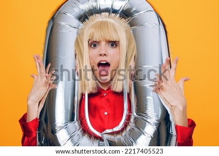 funny, woman stands on a yellow background in a red shirt and makes a funny face sticking out her tongue and holding a balloon in the form of the number zero in her hands, sticking her head into it