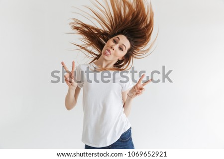 Funny woman in t-shirt showing peace gestures and her tongue while looking at the camera over grey background
