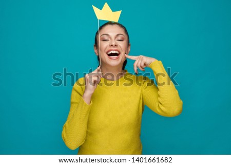 Funny woman in yellow sweater posing with paper crown on blue isolated background. Cheerful stylish girl laughing and relaxing in studio. Concept of  leisure and entertainment.