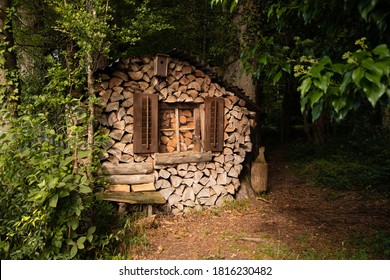 funny woodpile house in the middle of the green forest with a window and shutters and a bird house and a brown carnival mask with white teeth hanging in the window pane