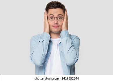 Funny worried man in glasses jean shirt hold head in hands looks concerned pose studio grey wall, concept of anxiety, irrevocable mistake, guy feels fear makes big eyes face expressions concept image
