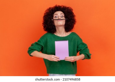 Funny young adult woman with Afro hairstyle wearing green sweater having fun holding pencil with lip and nose, female student resting during hometask. Indoor studio shot isolated on orange background.
