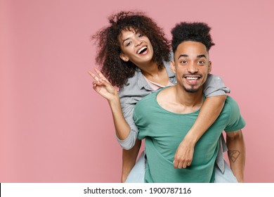 Funny young african american couple in love woman man in gray green casual clothes giving piggyback ride to joyful, sitting on back showing victory v sign isolated on pink background studio portrait