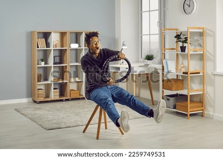 Funny, young African American man sitting on a chair in the living room at home, holding a black steering wheel cover in hands and pretending to drive an invisible car at a great speed