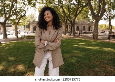 Funny Young African Woman Looks Away With Smile Spends Time In Park In Sunny Weather. Model With Brunette Wavy Hair Wears Jacket And White Jeans. Concept Lifestyle.