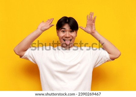 funny young Asian guy in a white T-shirt is grimacing and fooling around on a yellow isolated background, Korean man shows his tongue and makes grimaces