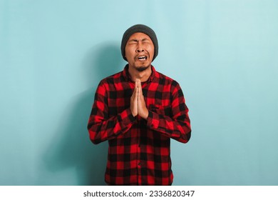 Funny Young Asian man with a beanie hat and red plaid flannel shirt is begging for forgiveness and praying with his hands together, isolated on a blue background