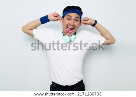 Funny young Asian sportman in blue headband and sportswear white t-shirt with headphones, hands fingers hold pull ears showing tongue out isolated on white background. Workout sport concept