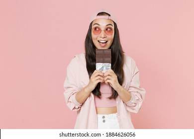 Funny Young Asian Woman 20s In Casual Clothes Cap Glasses Posing Isolated On Pastel Pink Wall Background Studio Portrait. People Emotions Lifestyle Concept. Mock Up Copy Space. Hold Chocolate Bar