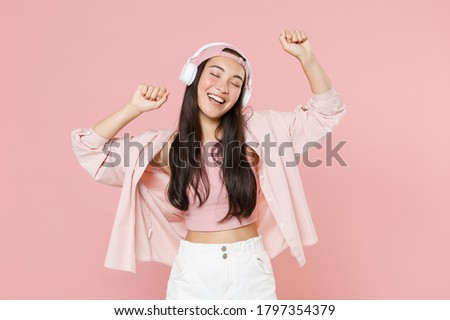 Funny young asian woman in casual clothes cap posing isolated on pastel pink background studio portrait. People lifestyle concept. Mock up copy space. Listening music with headphones, dancing
