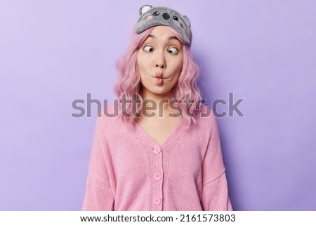 Funny young Asian woman grimaces at camera makes fish lips crosses eyes tries to make you laugh dressed in casual pink jumper wears blindfold on forehead mimicks isolated over purple background