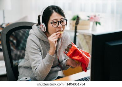 Funny Young Asian Woman Otaku Sit At Home With Pack Of Snacks In His Hands Eating Chips And Watching Videos On Computer Screen. Female Student On Summer Break Binge Watch On Internet With Junk Food.