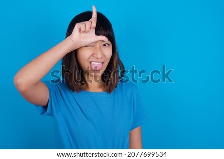 Funny young asian woman wearing blue t-shirt against blue background makes loser gesture mocking at someone sticks out tongue making grimace face.