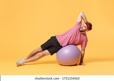 Funny young bearded fitness sporty guy 20s sportsman in headband t-shirt in home gym isolated on yellow background in studio. Workout sport motivation lifestyle concept. Doing side plank on fitball