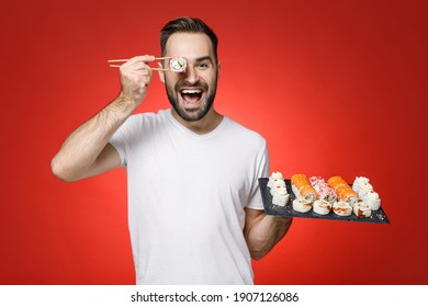 Funny Young Bearded Man 20s In Casual White T-shirt Covering Eye Hold Sticks Chopsticks Makizushi Sushi Roll Served On Black Plate Traditional Japanese Food Isolated On Red Background Studio Portrait