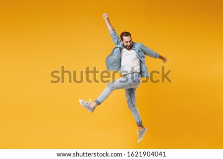 Funny young bearded man in casual blue shirt posing isolated on yellow orange wall background studio portrait. People sincere emotions lifestyle concept. Mock up copy space. Jumping, rising hands up