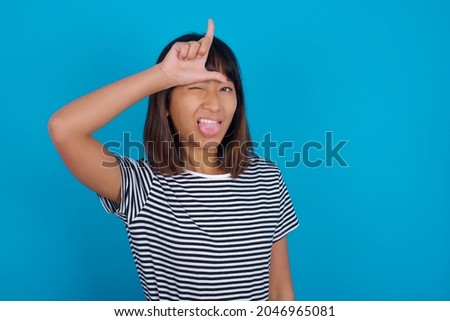 Funny Young beautiful asian girl wearing striped t-shirt over blue background makes loser gesture mocking at someone sticks out tongue making grimace face.