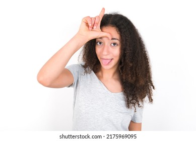 Funny Young Beautiful Brunette Teen Girl Wearing Grey T-shirt Over White Wall Makes Loser Gesture Mocking At Someone Sticks Out Tongue Making Grimace Face.