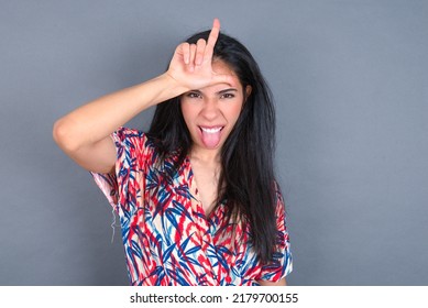 Funny Young Beautiful Brunette Woman Wearing Colourful Dress Over White Wall Makes Loser Gesture Mocking At Someone Sticks Out Tongue Making Grimace Face.