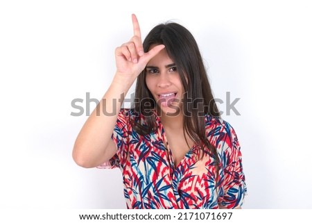Funny young beautiful brunette woman wearing colourful dress over white wall makes loser gesture mocking at someone sticks out tongue making grimace face.
