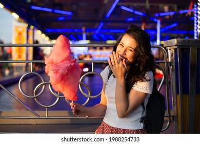 Funny young beautiful woman eating cotton candy at fairground at sunset