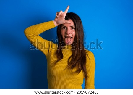 Funny young beautiful woman wearing yellow sweater over blue studio background makes loser gesture mocking at someone sticks out tongue making grimace face.