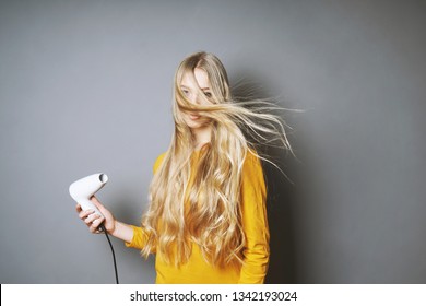 Funny Young Blond Woman Blow-drying Her Long Hair With Blow Dryer