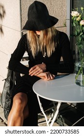 Funny Young Blond Woman Pulled A Floppy Hat Over Her Face. She Is Wearing Long Sleeved Balck Dress, Sitting Behind Cafe Table Outside.