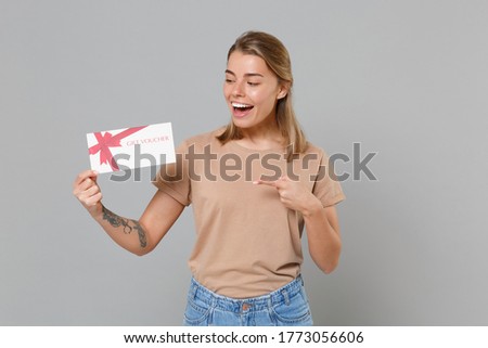 Funny young blonde woman girl in casual beige t-shirt posing isolated on gray wall background studio portrait. People lifestyle concept. Mock up copy space. Pointing index finger on gift certificate