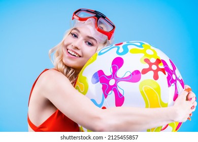 Funny young blonde woman in red swimsuit and goggles is posing, holding ball, isolated on blue background. Emotional girl ready to swim and play beach games. Summer vacation rest lifestyle concept. 