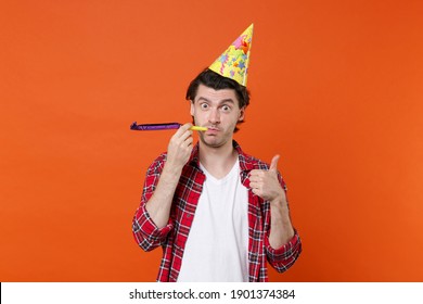Funny young brunet man 20s in casual clothes white t-shirt red checkered shirt birthday hat posing blowing in pipe showing thumb up looking camera isolated on orange color background studio portrait - Shutterstock ID 1901374384
