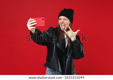 Funny young brunette woman 20s in casual black leather jacket white t-shirt hat doing selfie shot on mobile phone depicting heavy metal rock sign isolated on bright red background studio portrait