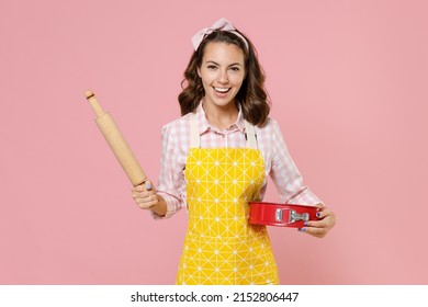 Funny Young Brunette Woman Housewife 20s Wearing Yellow Apron Hold Rolling Pin Metal Baking Form For Pie While Doing Housework Isolated On Pastel Pink Background Studio Portrait. Housekeeping Concept