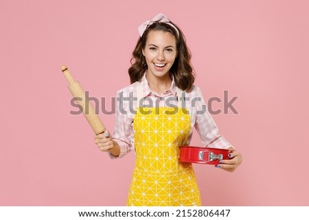 Funny young brunette woman housewife 20s wearing yellow apron hold rolling pin metal baking form for pie while doing housework isolated on pastel pink background studio portrait. Housekeeping concept