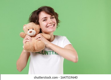 Funny young brunette woman in white volunteer t-shirt isolated on pastel green background in studio. Voluntary free work assistance help charity grace teamwork concept. Holding teddy bear plush toy