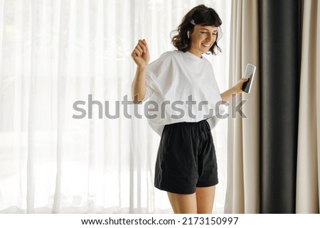 Funny young caucasian girl dances to music listened through headphones in light room. Brunette wears white t-shirt and black shorts. Morning holiday concept