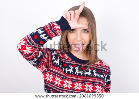 Funny Young caucasian girl wearing christmas sweaters on white background makes loser gesture mocking at someone sticks out tongue making grimace face.