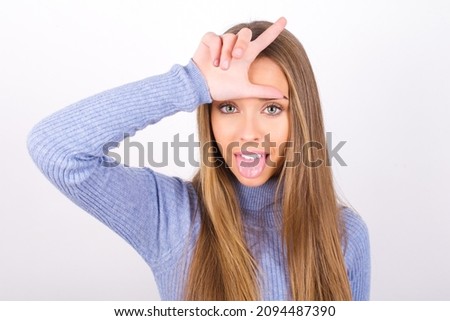 Funny Young caucasian girl wearing blue turtleneck over white background makes loser gesture mocking at someone sticks out tongue making grimace face.